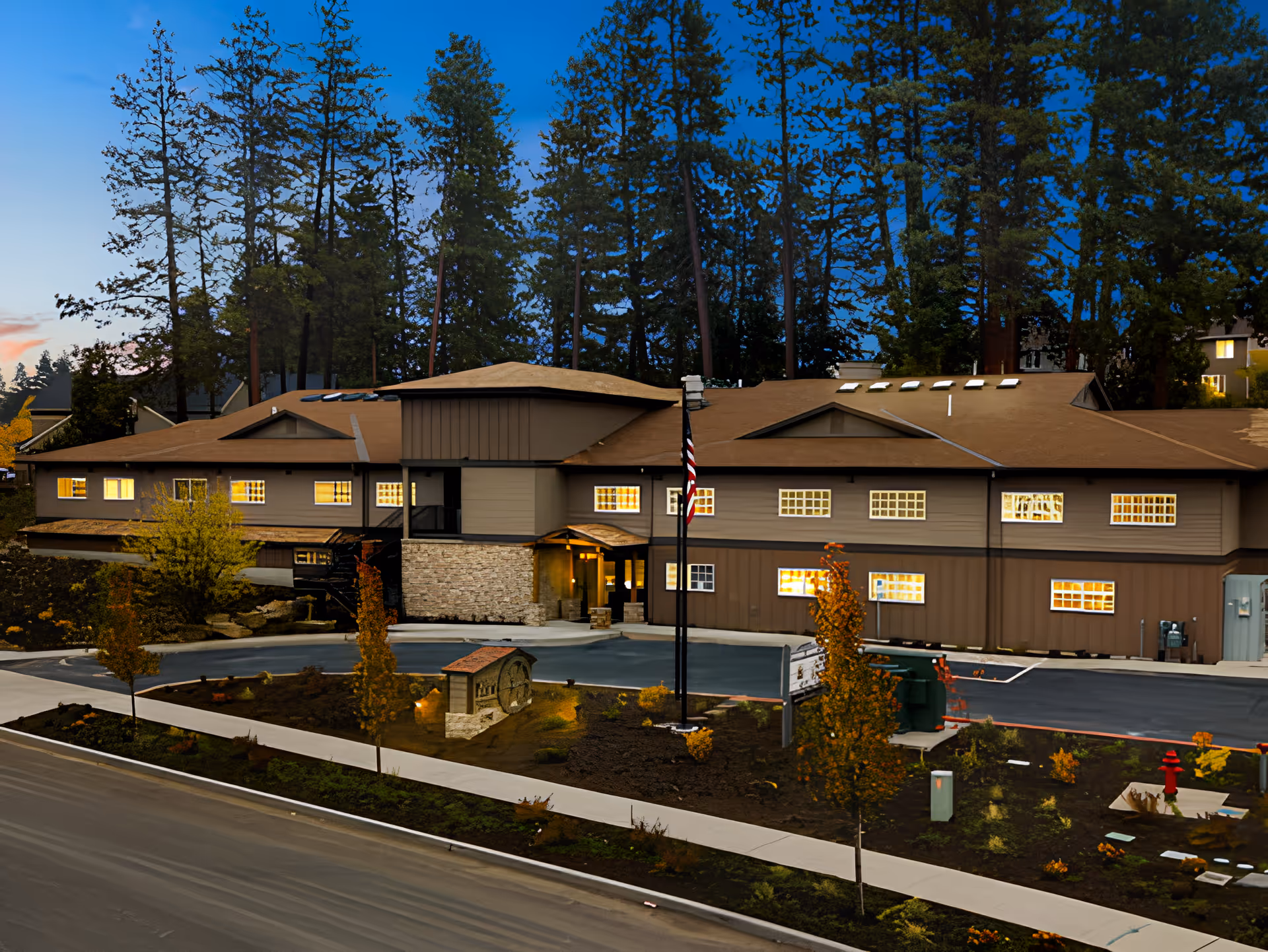 Exterior view of Mill View Memory Care facility at dusk, showing a two-story building with illuminated windows, surrounded by tall trees and landscaped grounds with small trees and shrubs. An American flag is displayed near the entrance.