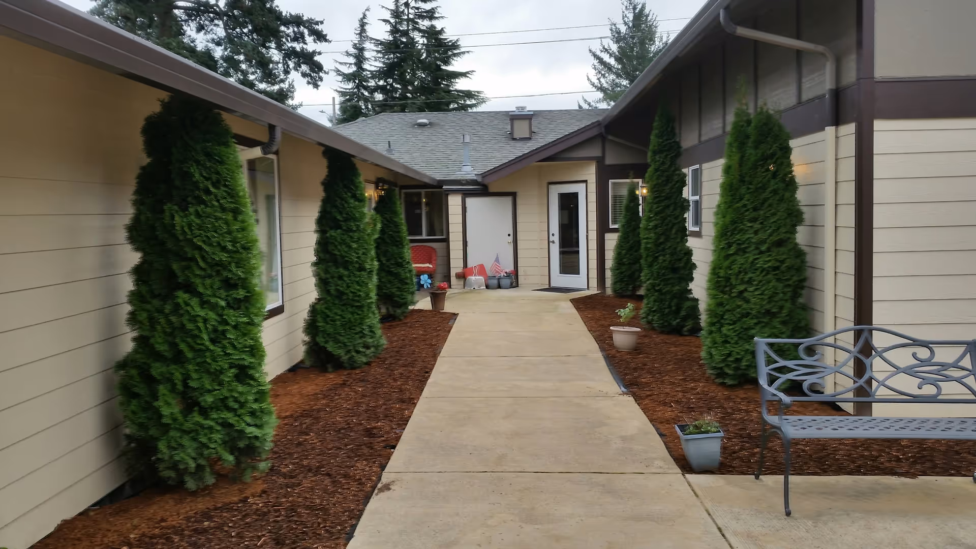Outdoor walkway between two beige buildings with brown trim, lined with tall green shrubs and small potted plants. A metal bench is placed on the right side near the walkway. The sky is overcast and tall trees are visible in the background.