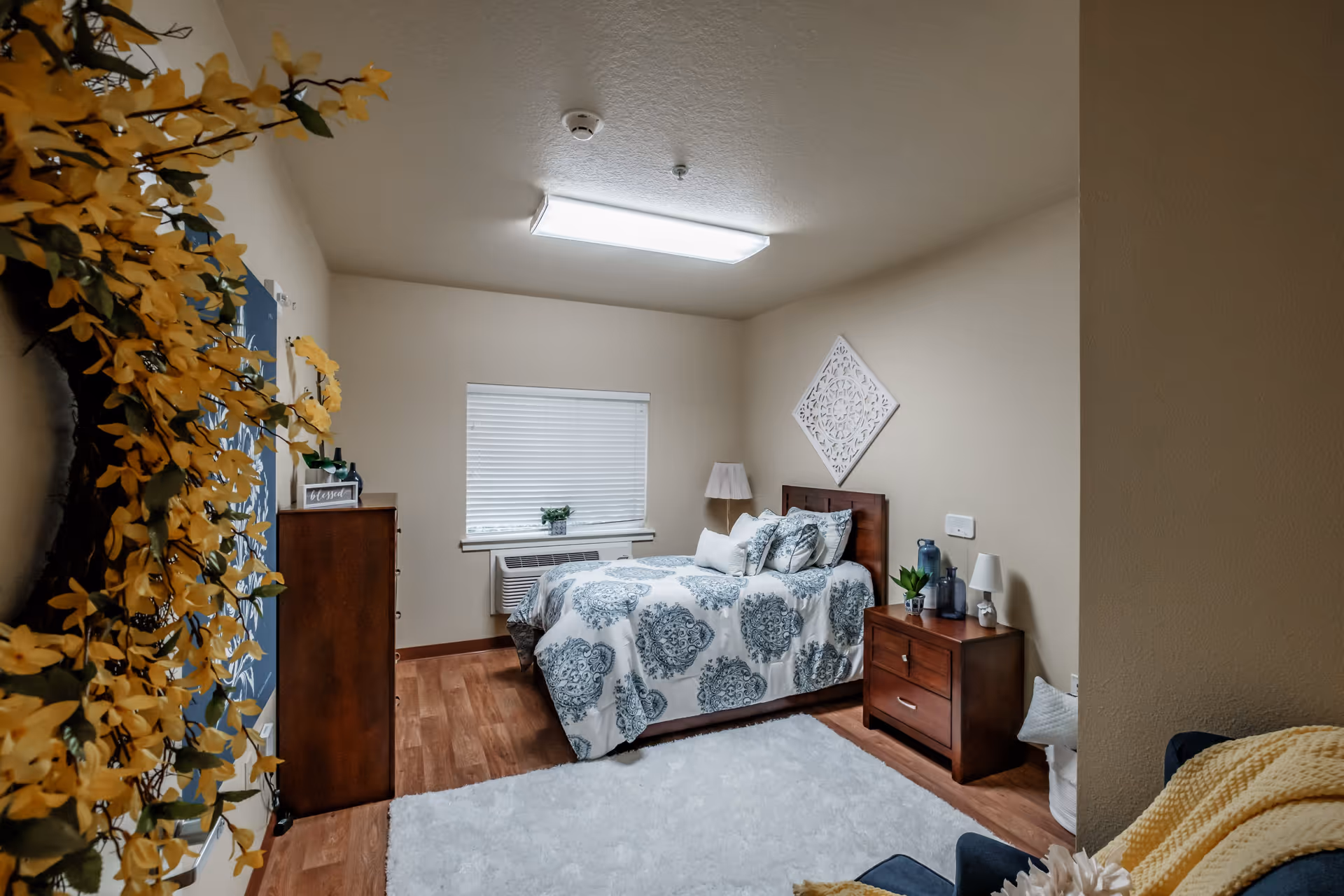A cozy bedroom with a single bed covered in a white and blue patterned comforter and multiple pillows. The room has wooden flooring, a white fluffy rug, a wooden nightstand with decorative items and a lamp, a wooden dresser, a window with blinds, and a wall decoration above the bed. Yellow flowers are partially visible on the left side of the image.