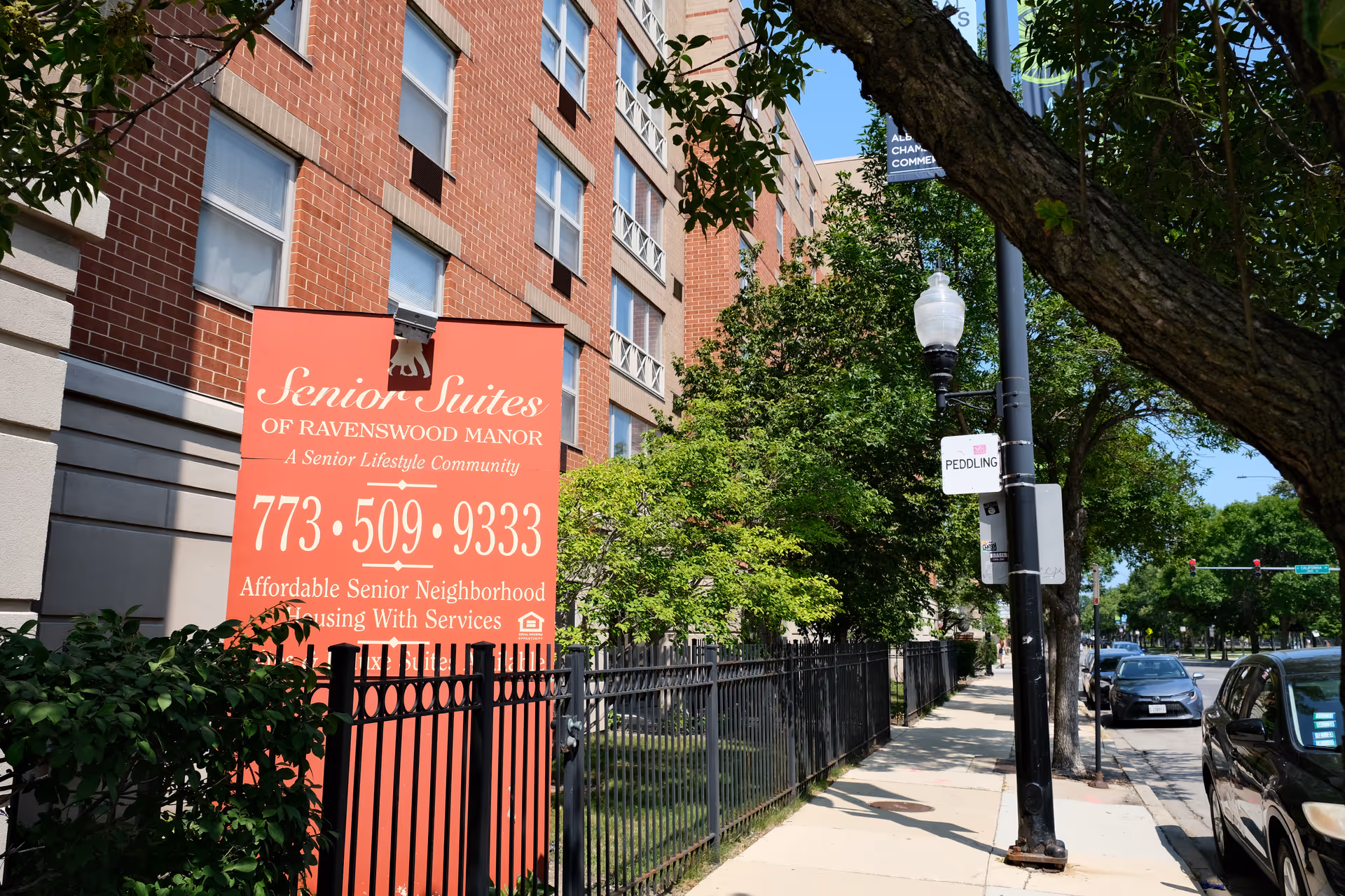 Exterior view of a multi-story brick building with a large red sign in front that reads 'Senior Suites of Ravenswood Manor, A Senior Lifestyle Community, 773-509-9333, Affordable Senior Neighborhood Housing With Services'. The building is surrounded by trees, a black metal fence, and a sidewalk with parked cars and a street lamp.