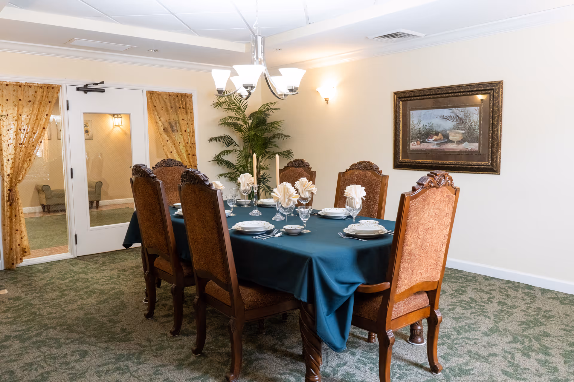 A formal dining room with a rectangular table covered by a dark green tablecloth, set with plates, glasses, and folded napkins. Six ornate wooden chairs surround the table. The room has patterned green carpet, cream-colored walls, a framed painting on the wall, a potted plant in the corner, and a chandelier overhead. There is a door with glass panels and sheer gold curtains on the left side.