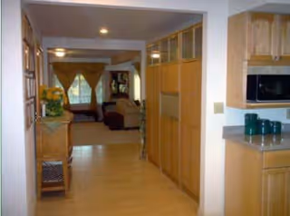 View down a hallway with light wood flooring and cabinetry on the right side, leading to a living room area with a sofa and large windows with curtains. A small table with a plant is on the left side of the hallway.