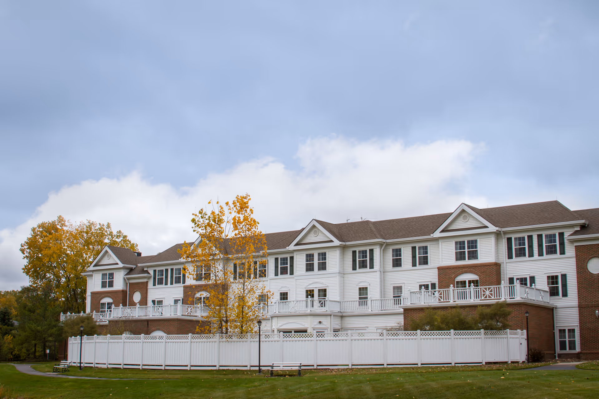 Exterior view of a large, multi-story senior living facility building with white siding and brick accents. The building has multiple windows and balconies with white railings. There is a white fence surrounding a portion of the property, green grass, and trees with autumn foliage. The sky is cloudy.