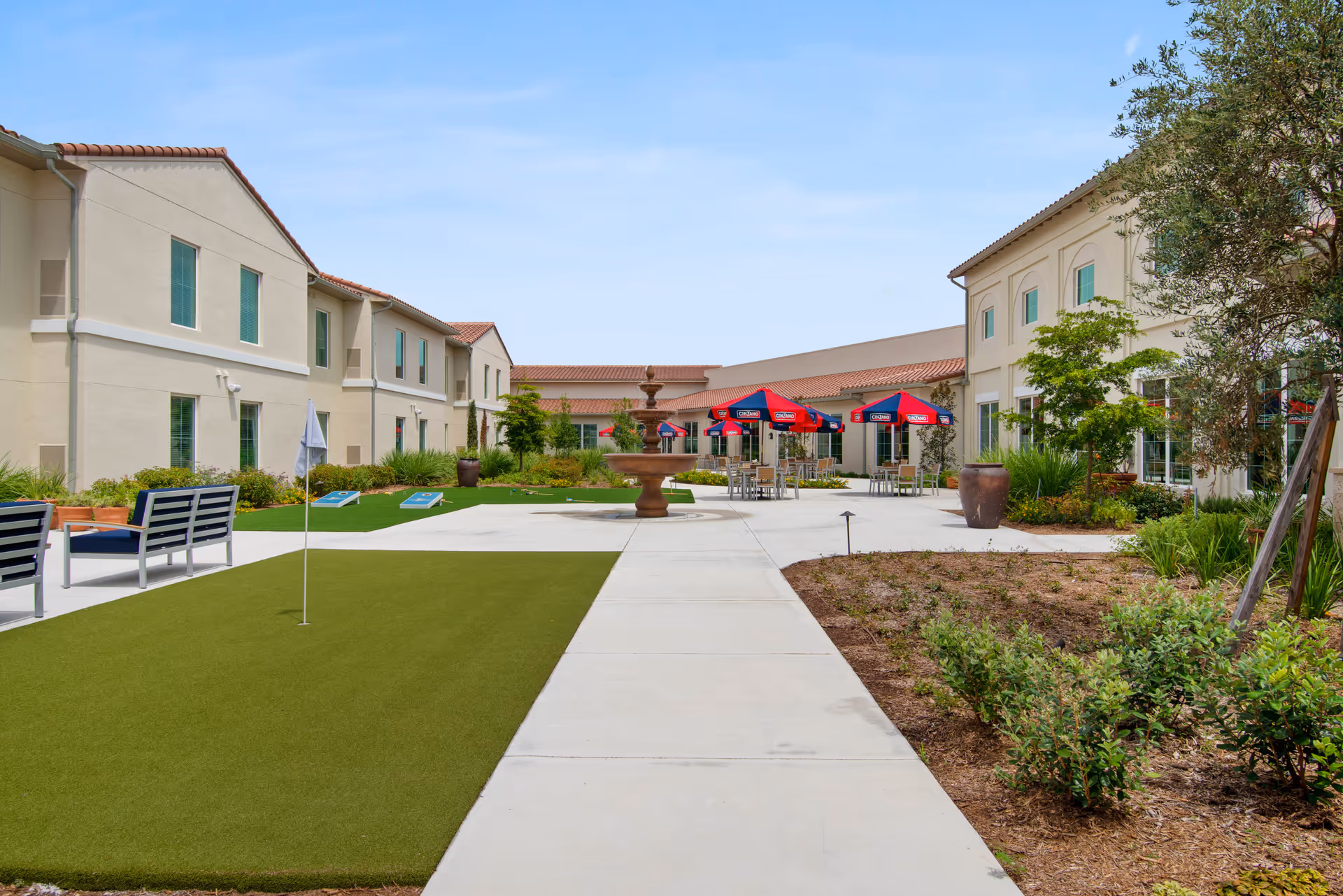 Sunny courtyard of a senior living facility with a central fountain, seating, umbrellas, a putting green, and surrounding two-story buildings.