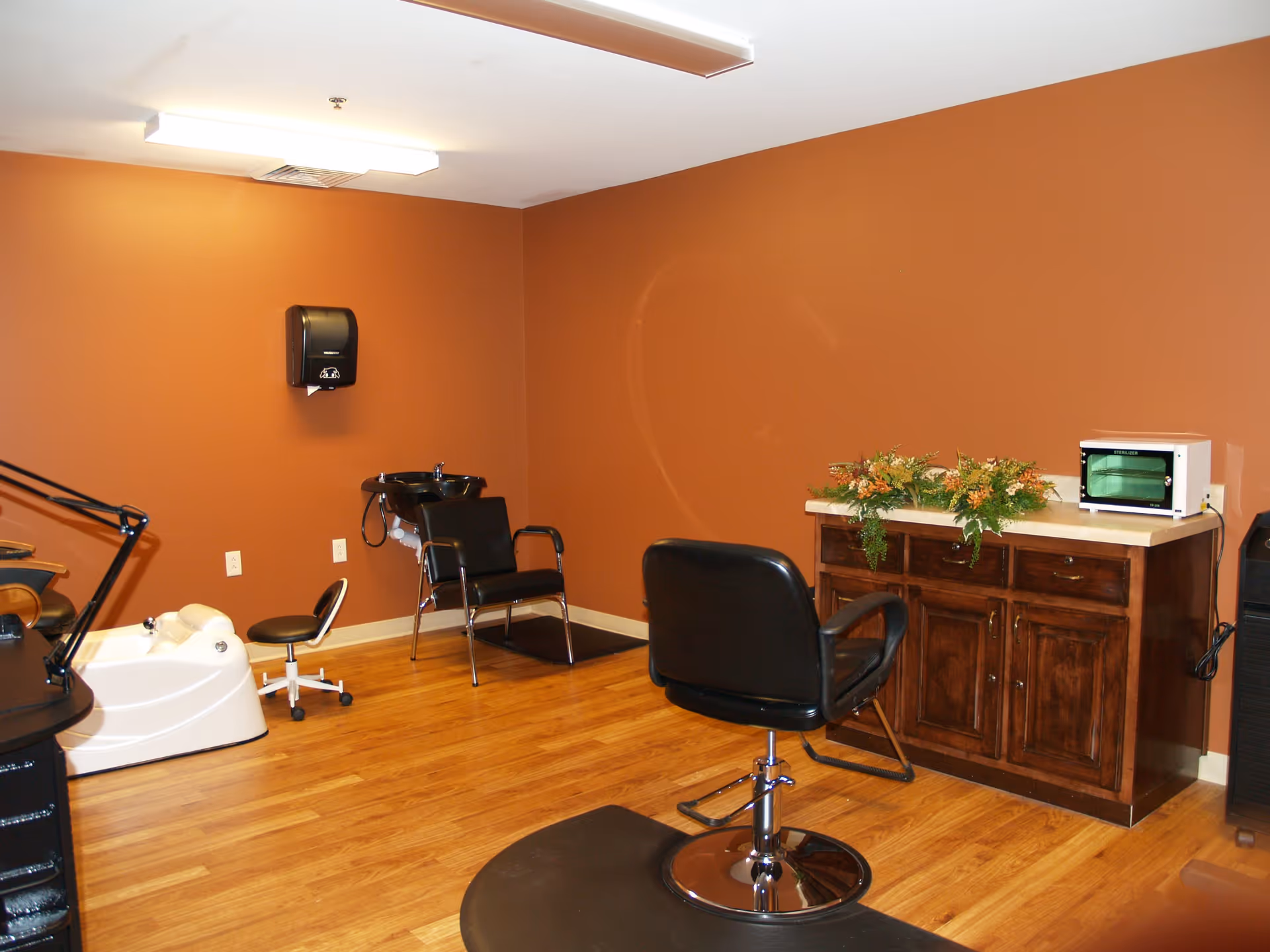 Small salon room with styling chairs, a shampoo sink and pedicure tub next to a wooden cabinet against brown walls and hardwood floors.