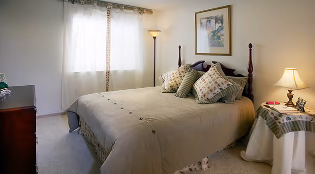 A neatly made bedroom with a double bed, decorative pillows, bedside tables with lamps, a dresser, and a curtained window.