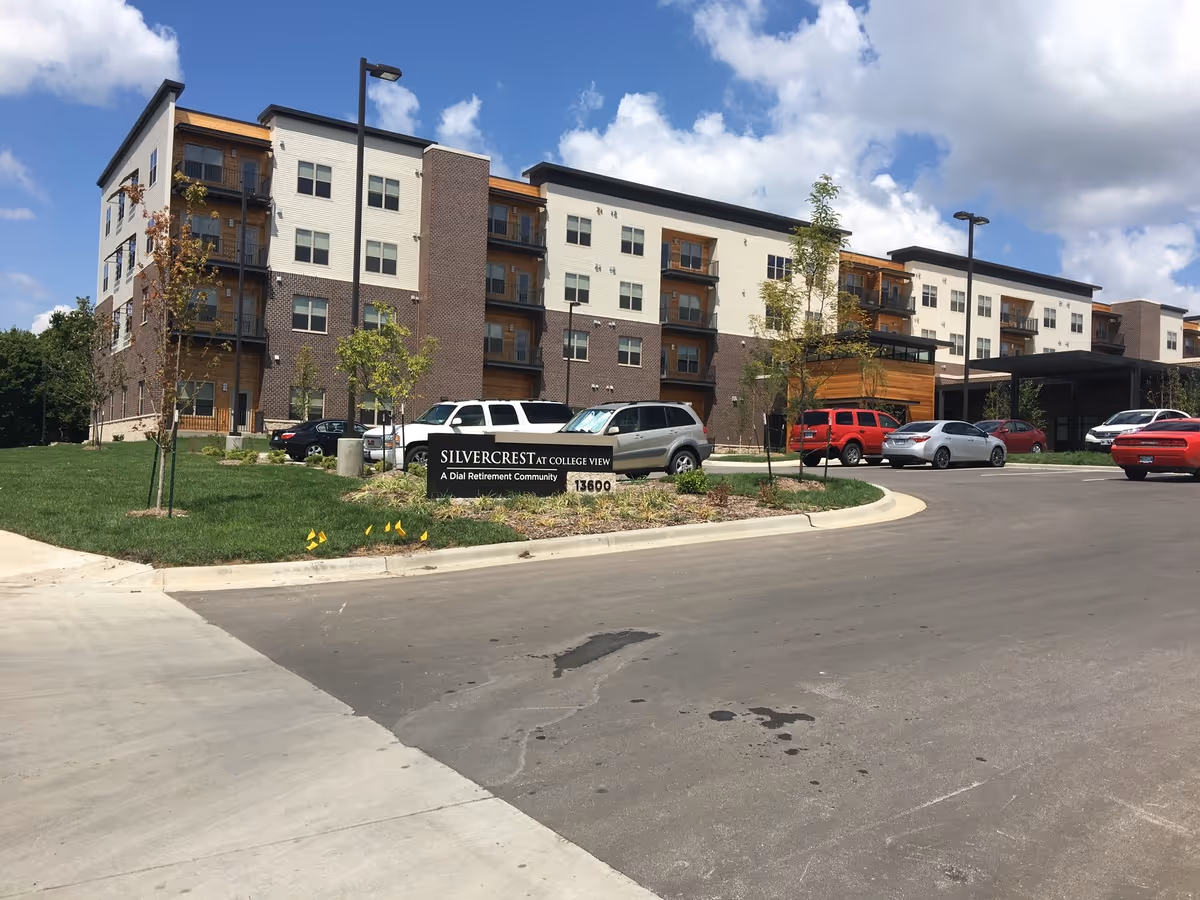 Exterior view of a modern multi-story senior living facility named Silvercrest at College View with a parking lot in front, several cars parked, young trees planted on a grassy area, and a partly cloudy sky.