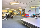 A spacious physical therapy or rehabilitation room with exercise equipment including stationary bikes, parallel bars, and therapy beds. The room has carpeted flooring, fluorescent ceiling lights, and a door at the far end. There are some therapy tools and a small red and blue children's ride-on toy visible.