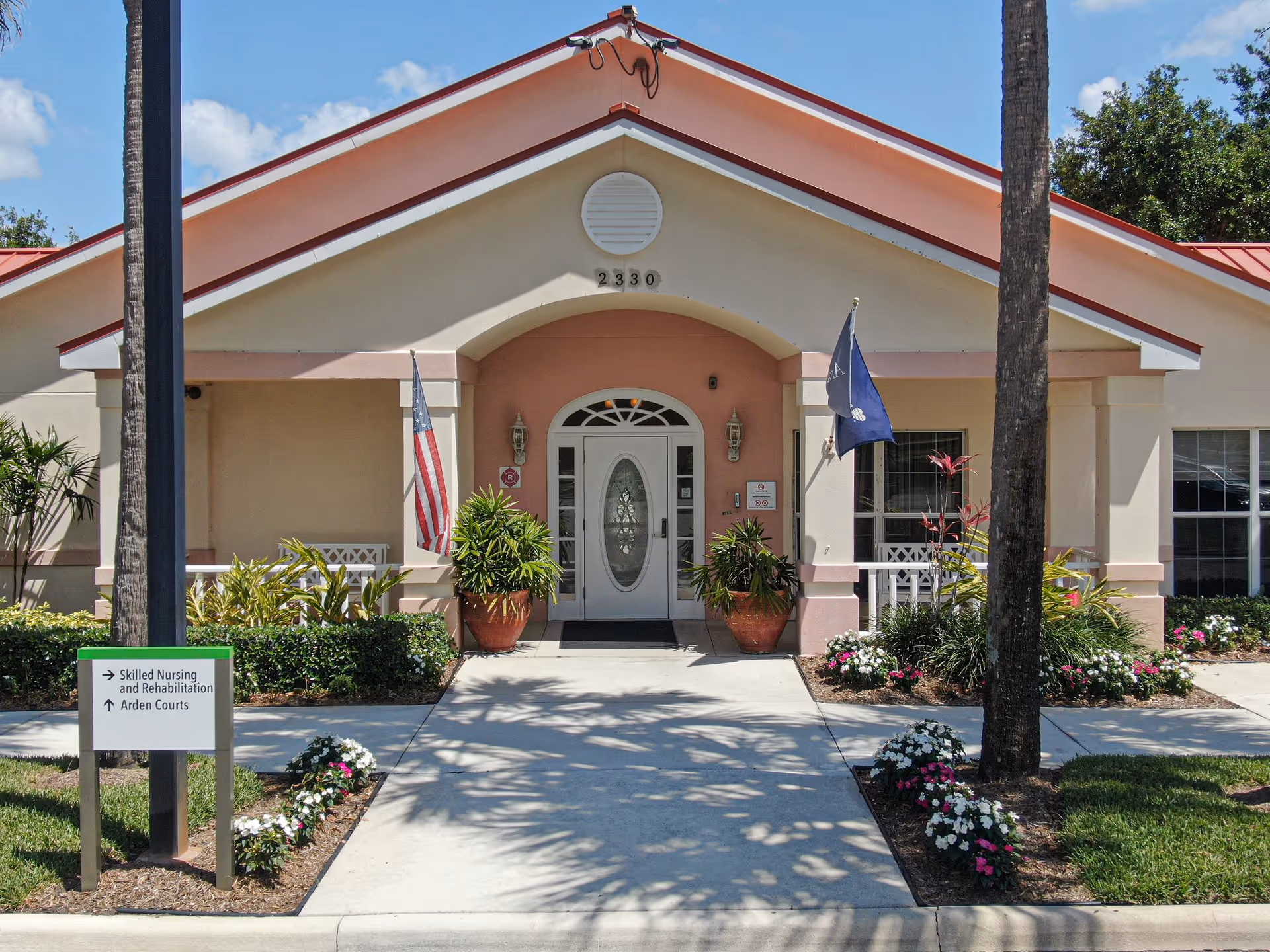 Front exterior view of Arden Courts - ProMedica Memory Care Community building with a walkway leading to a white door entrance. The building has a peach and cream color scheme, two potted plants flanking the door, an American flag and another flag on poles, and a sign indicating directions to Skilled Nursing and Rehabilitation and Arden Courts. Palm trees and flower beds line the walkway.
