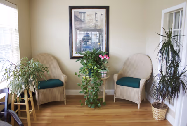 Two wicker chairs with green cushions flank a small table topped with plants beneath a framed picture in a sunlit sitting area.
