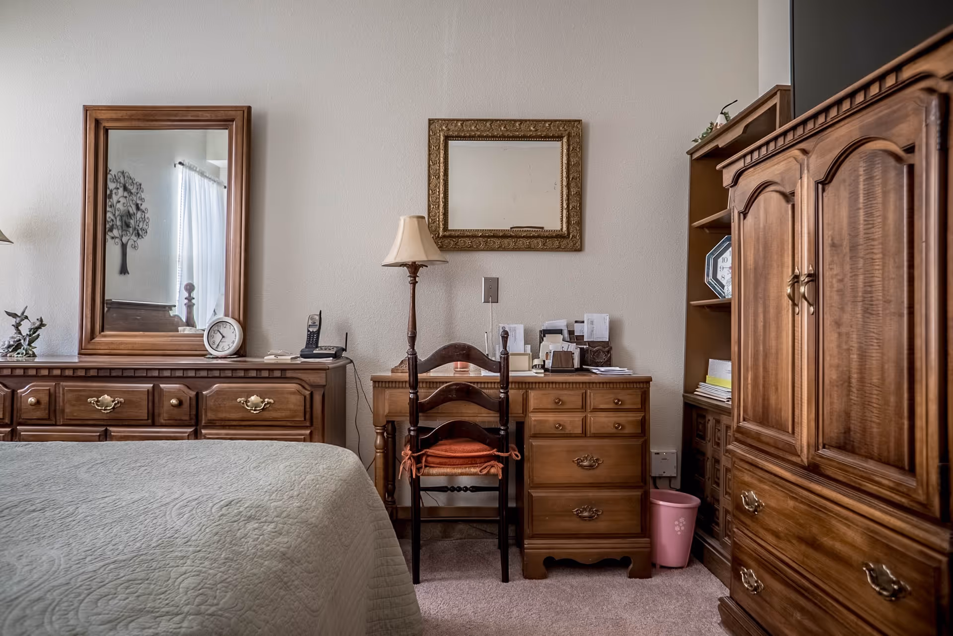 A bedroom with wooden furniture including a dresser with a mirror, a desk with a chair, a tall cabinet, and a bookshelf. The bed with a light-colored quilt is partially visible on the left. A table lamp and a clock are on the dresser, and a framed mirror hangs on the wall above the desk.