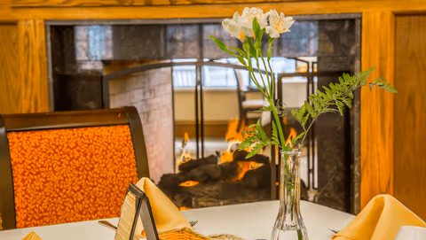 Dining table set with folded napkins and a vase of flowers in front of a lit fireplace and a chair.