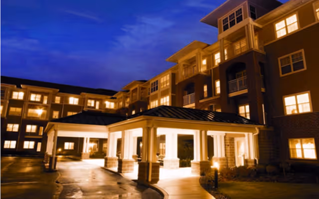 Nighttime exterior of a multi-story senior living building with a lit covered entrance and many illuminated windows.