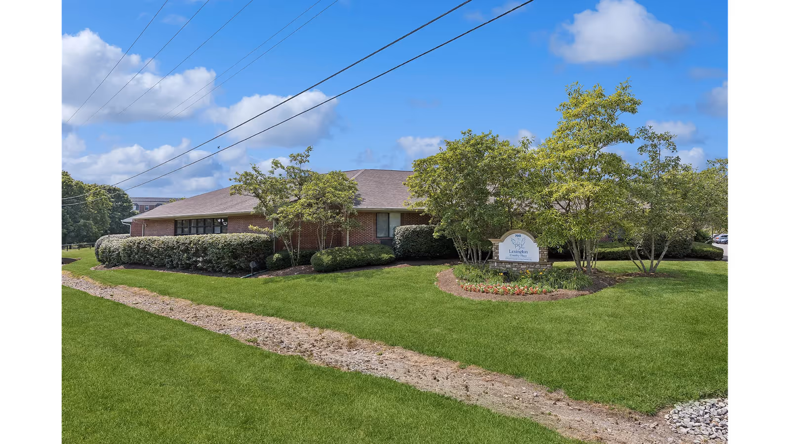 Single-story brick building with a sloped roof surrounded by green grass, bushes, and trees under a blue sky with some clouds. A sign in front of the building reads 'Lexington Living Place.'
