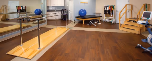 A rehabilitation or physical therapy room with wooden flooring, parallel bars for walking exercises, a blue exercise ball, a small set of stairs with handrails, exercise equipment, and a large mirror on one wall.