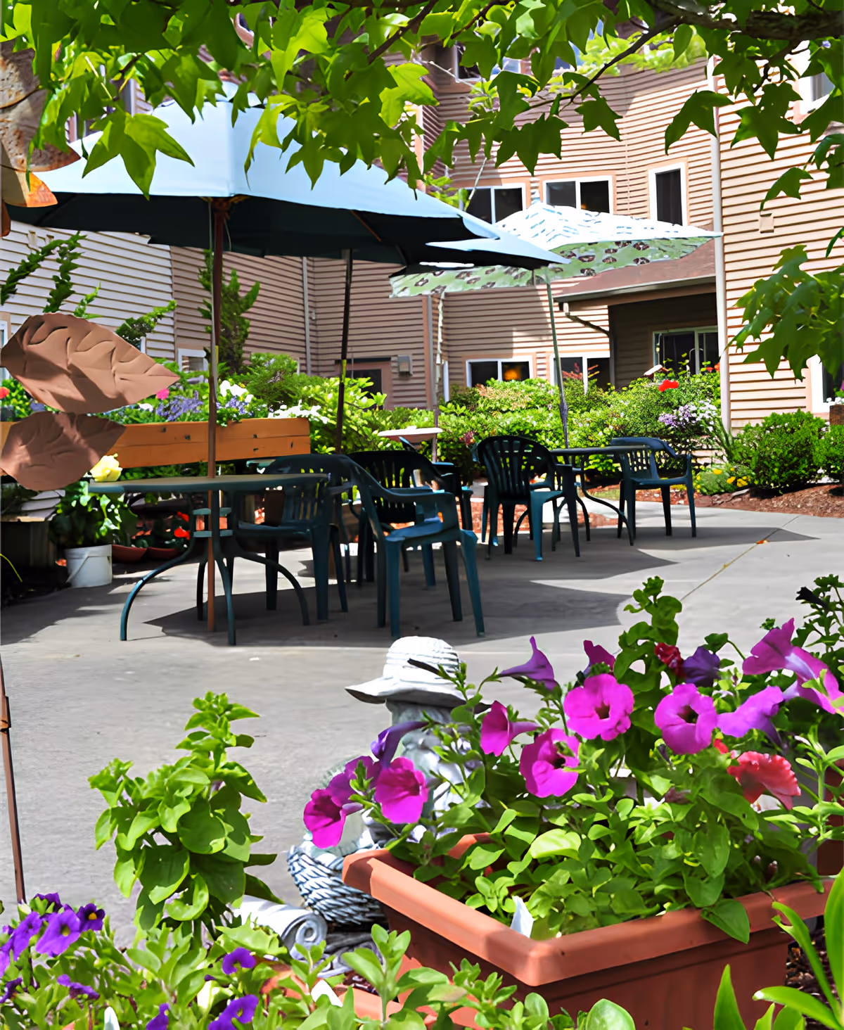 Outdoor patio area at Vineyard Park of Port Orchard with several tables and chairs under large umbrellas. The patio is surrounded by lush greenery and colorful flowers, with a building visible in the background.