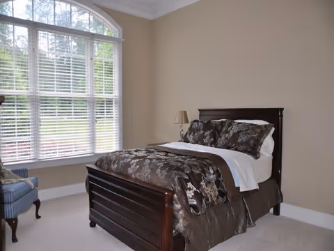 A bedroom with a large wooden bed featuring dark floral bedding and white sheets. There is a bedside table with a lamp on the right side of the bed. A large window with white blinds allows natural light to fill the room. A blue upholstered chair is positioned near the window. The walls are painted beige and the floor is carpeted in a light color.