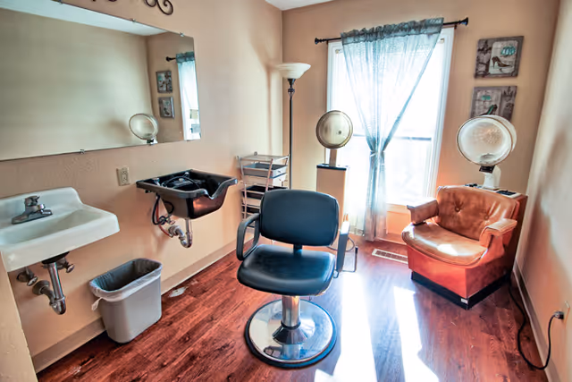 A small salon room with a black salon chair in the center, a black hair washing sink attached to the wall, a white sink with a faucet, a trash bin underneath, a brown leather armchair, two vintage hair dryers on stands, a tall floor lamp, and a window with sheer blue curtains letting in sunlight.