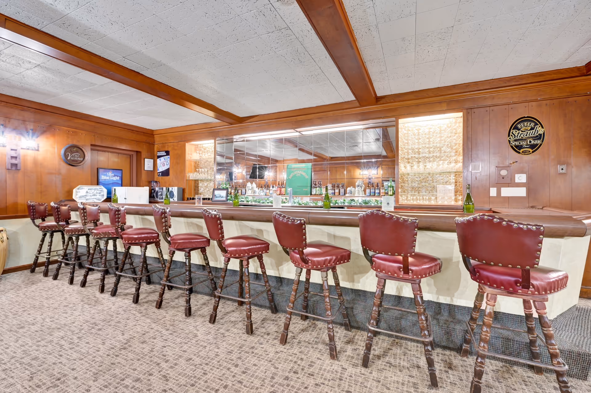 Interior view of a bar area with a long counter and nine high wooden bar stools with red cushioned seats and backs. The bar has a mirrored back wall with shelves holding various bottles and glasses. The walls are wood-paneled, and there are decorative signs and lighting fixtures on the walls. The ceiling has white tiles with wooden beams.