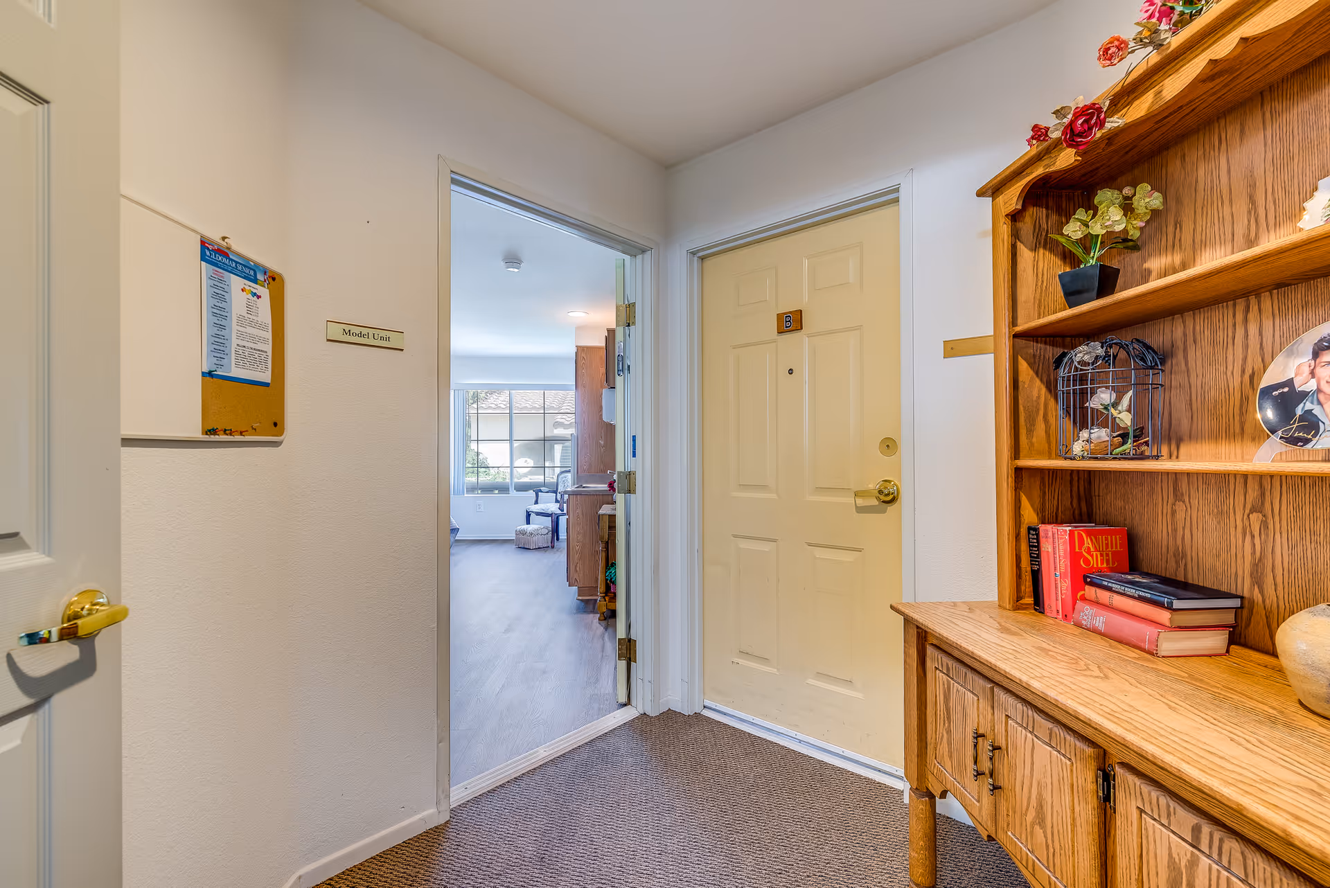 Entry hallway of a model senior assisted living unit with a closed front door, open doorway into a living room, and a wooden bookshelf with books and decor.
