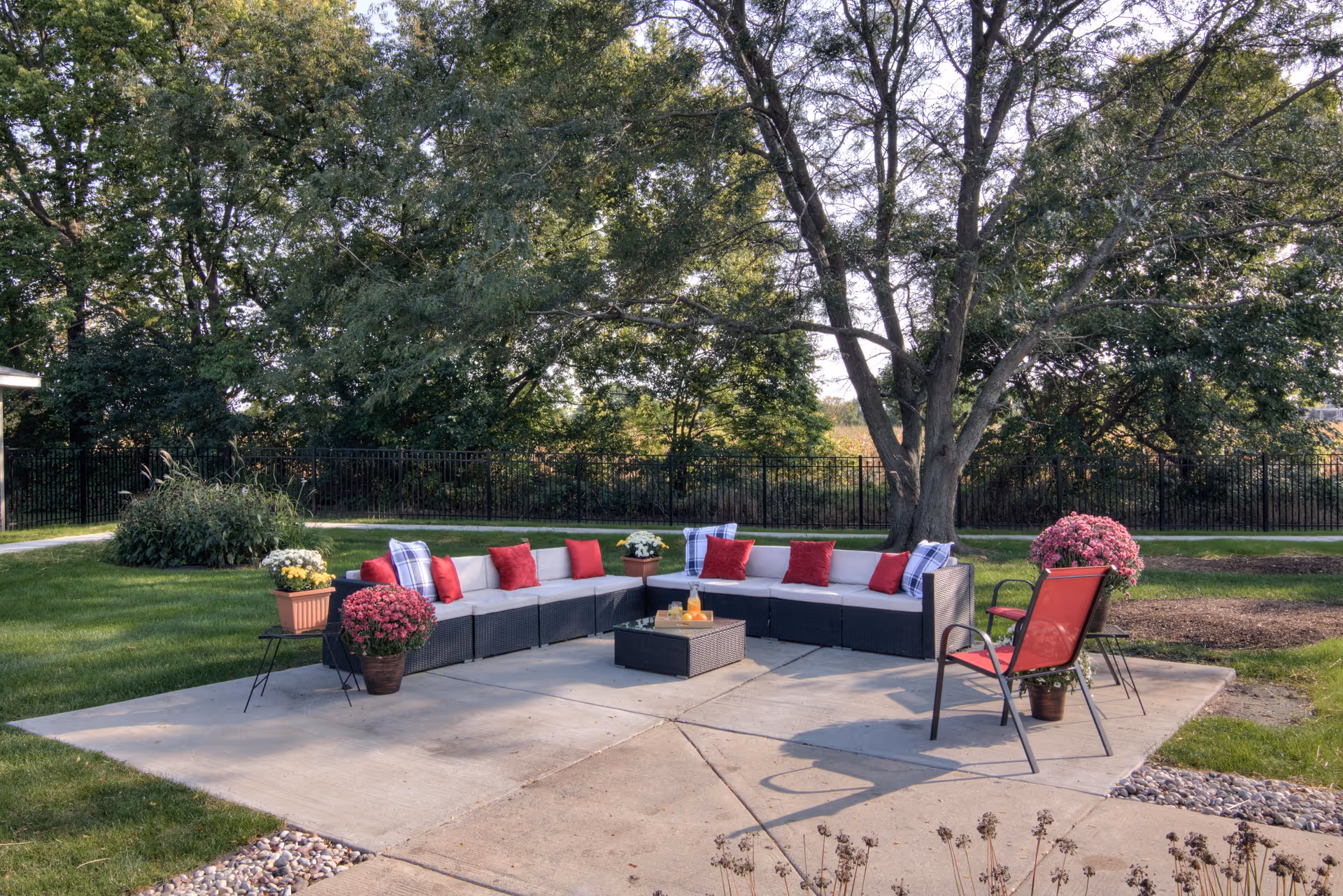 Outdoor seating area with a large L-shaped sectional sofa adorned with red and plaid pillows, a small square table in the center with drinks, two red chairs, and several potted plants and flowers. The area is surrounded by green grass, trees, and a black metal fence in the background.