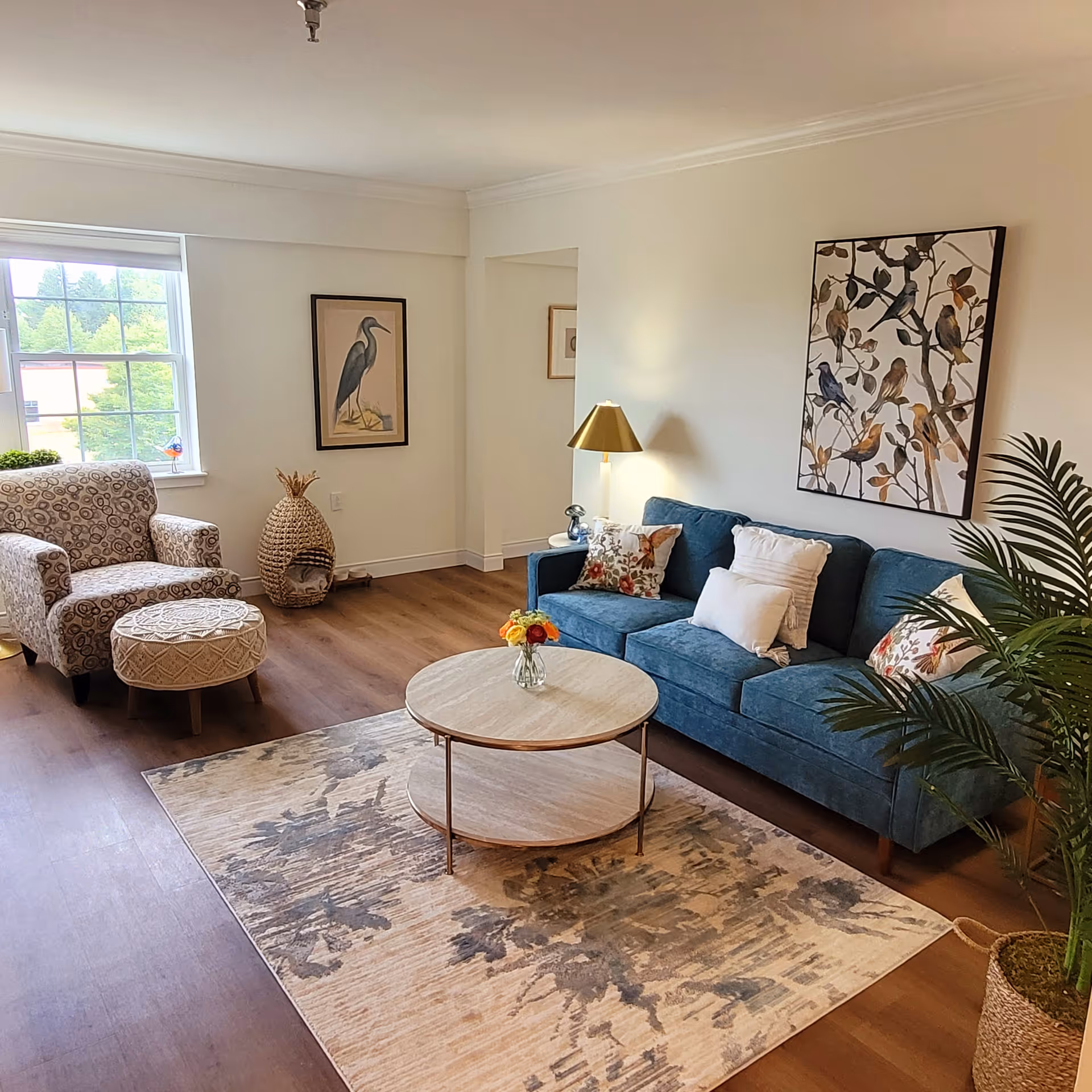 A cozy living room with a blue sofa adorned with decorative pillows, a round wooden coffee table with a small vase of flowers, a patterned armchair with a matching ottoman, a large window letting in natural light, framed bird artwork on the walls, a floor lamp with a gold shade, and a potted plant in a woven basket.
