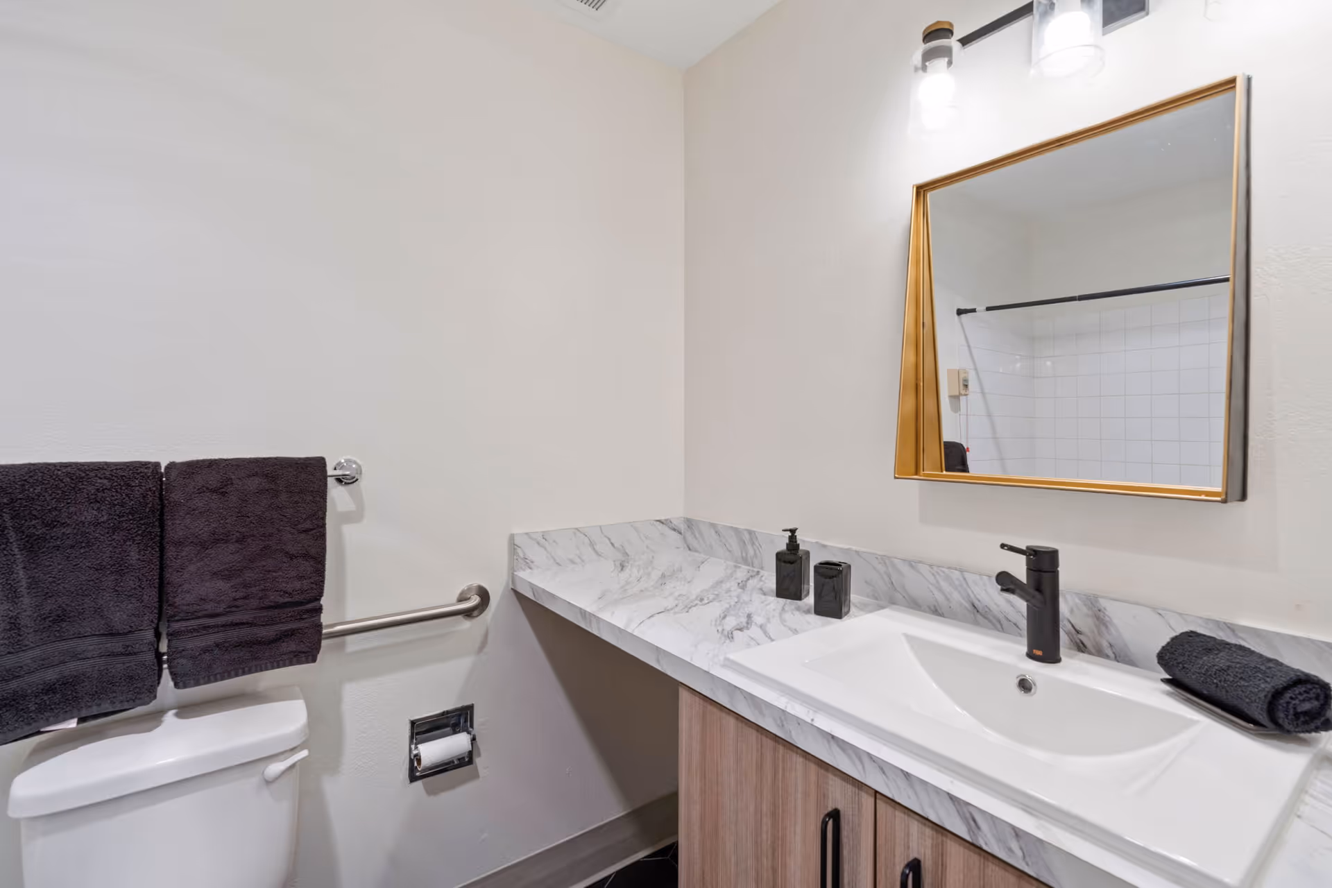 A clean bathroom with a white toilet, two dark towels hanging on a silver towel bar, a marble countertop with a white sink, black soap dispenser and toothbrush holder, a rolled dark towel, and a rectangular mirror with a gold frame above the sink. White tiled shower area is partially visible in the mirror reflection.