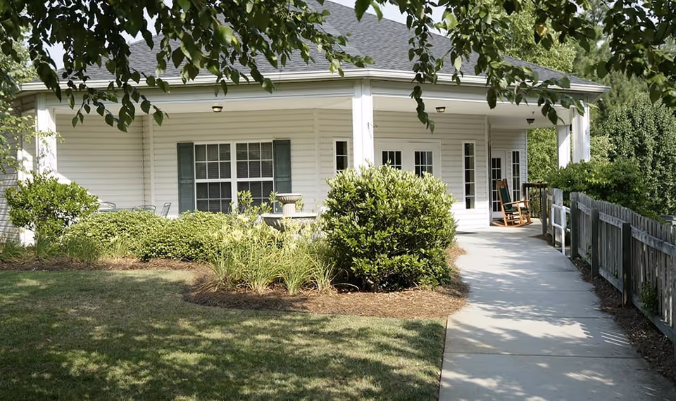 A covered porch area of a senior living facility with white siding, green shutters, and a concrete walkway leading to the entrance. There are bushes and plants along the walkway and a wooden rocking chair on the porch. Trees provide shade over the scene.