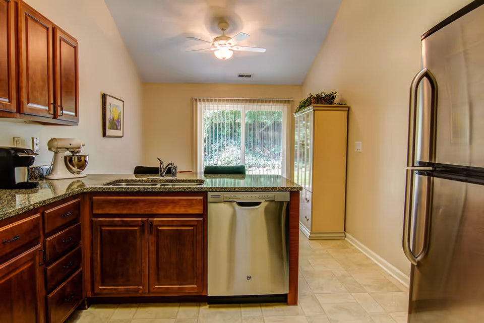 A kitchen with wooden cabinets, granite countertops, a stainless steel dishwasher, and a refrigerator. There is a sink with a faucet on the countertop, a coffee maker, and a stand mixer. A ceiling fan with a light is mounted on the ceiling, and a large window with vertical blinds overlooks a green outdoor area. A cabinet with plants on top is visible in the background.