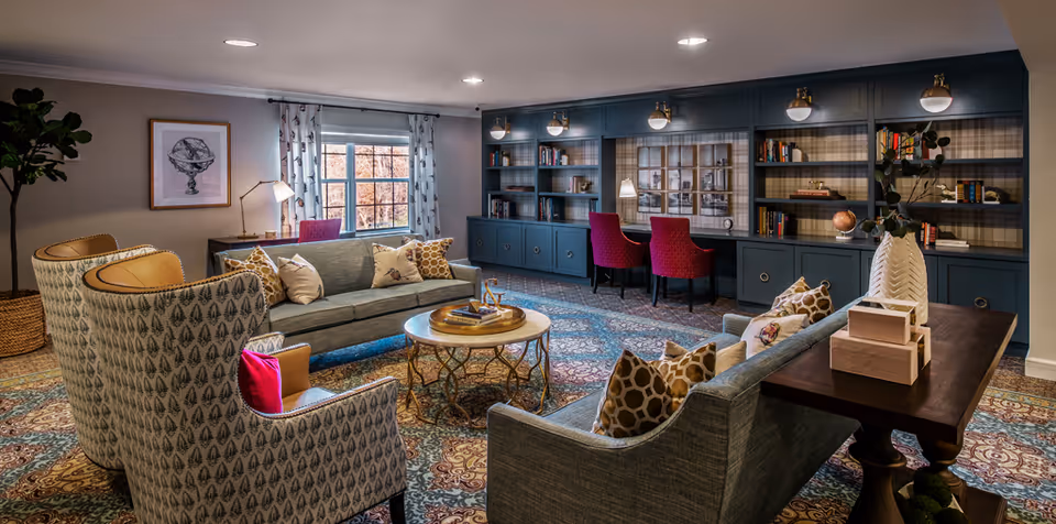 A cozy and well-decorated living room area with two patterned armchairs, two gray sofas with decorative pillows, a round coffee table, and a wooden console table with decorative items. The room features a large window with curtains, a wall with built-in blue bookshelves and cabinets, and two red upholstered chairs at a desk area. The carpet has an intricate pattern, and the lighting includes ceiling lights and wall-mounted lamps.