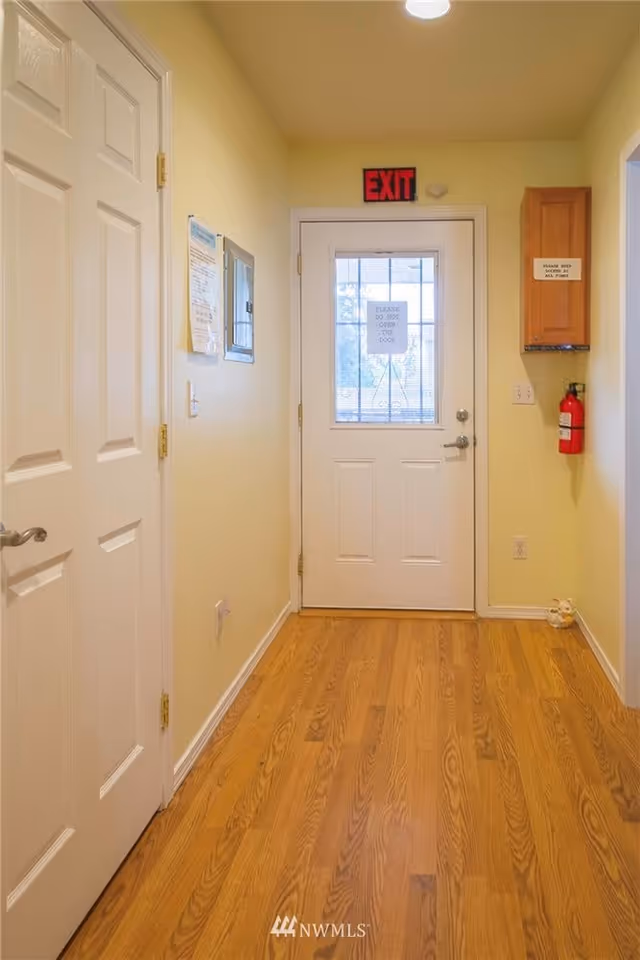 A hallway with light yellow walls and wooden flooring leading to a white door with a window and an exit sign above it. There is a fire extinguisher mounted on the right wall below a wooden cabinet, and a sign on the door reads 'PLEASE DO NOT OPEN THIS DOOR'. On the left wall, there are some papers and a small electrical panel.