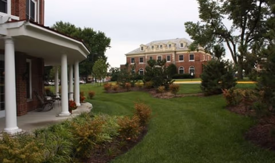 View of a landscaped outdoor area at Armour Oaks Senior Living Community featuring a green lawn, shrubs, and trees. On the left side, there is a covered porch with white columns and chairs. In the background, a large brick building with multiple windows and a light-colored roof is visible under a cloudy sky.