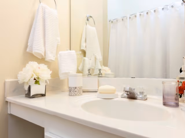 A clean bathroom countertop with a sink, a bar of soap on a soap dish, a toothbrush holder, a purple cup, a small vase with white flowers, and white towels hanging on a towel ring and rack. A white shower curtain is visible in the background.