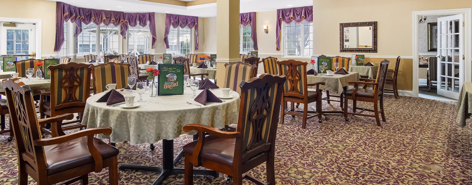 Bright dining room with round tables set for meals, wooden upholstered chairs, patterned carpet and purple window valances.