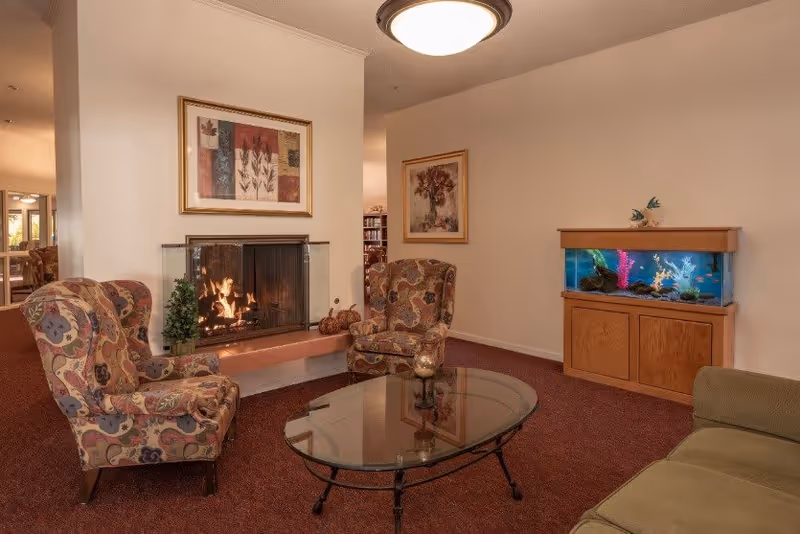 A cozy senior living room with two patterned armchairs, a glass coffee table, a green sofa, a fireplace with a fire burning, framed artwork on the walls, and a wooden cabinet with a fish tank on top.