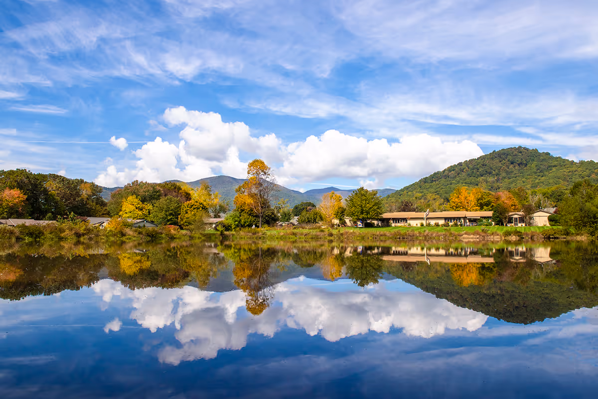 Low buildings and trees along a lakeshore reflected in calm water with blue sky, clouds, and distant hills.