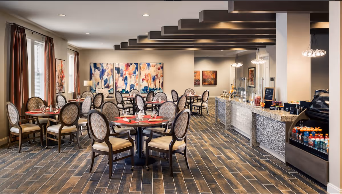 Bright, modern dining room with round tables and upholstered chairs, a buffet counter with drinks, and colorful artwork on the walls.