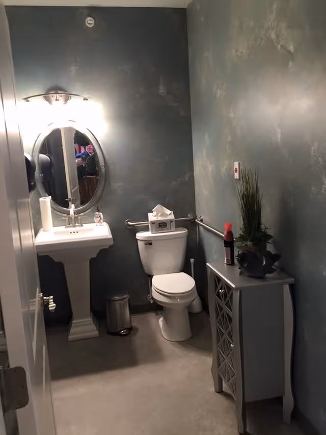 A small bathroom with a white pedestal sink, an oval mirror above it, and a light fixture on the wall. There is a white toilet with a tissue box on top, a small silver trash can on the floor, and a gray cabinet with decorative lattice doors holding a plant and a spray bottle. The walls have a textured blue-gray finish and there are metal grab bars installed near the toilet.