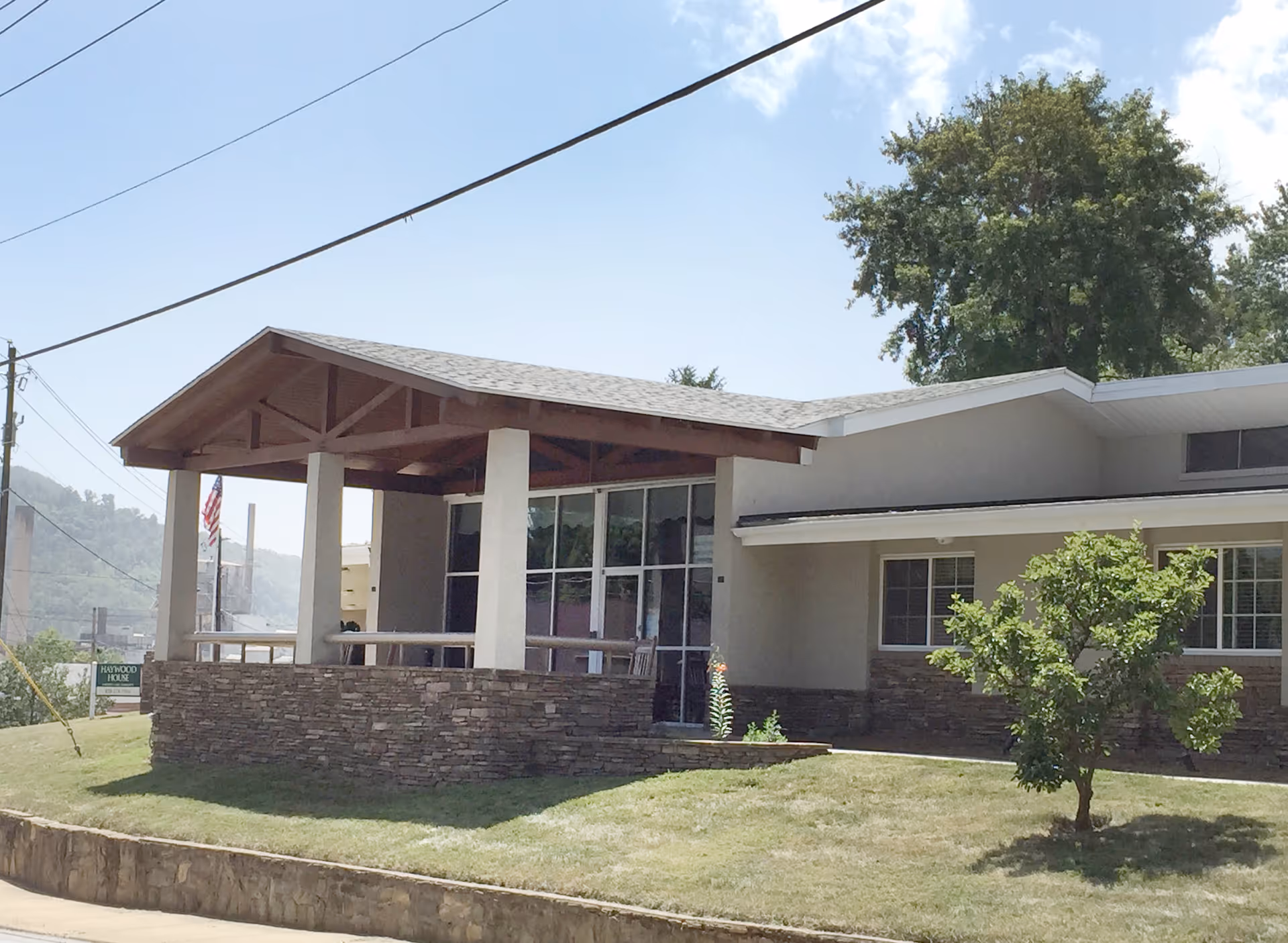 Exterior view of Haywood House, a single-story building with a covered porch supported by white pillars and a stone base. There is a small tree and grassy lawn in front, with a clear blue sky and some power lines overhead.