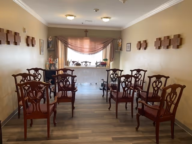 Rows of wooden chairs arranged facing a draped window and small altar-like display in a senior living common room with wood flooring.