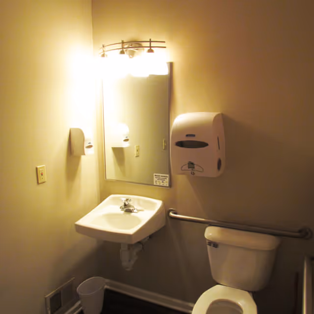 A small bathroom with a white sink mounted on the wall, a mirror above the sink with a light fixture, a white toilet with a grab bar next to it, a wall-mounted soap or paper towel dispenser, and a small trash bin on the floor.