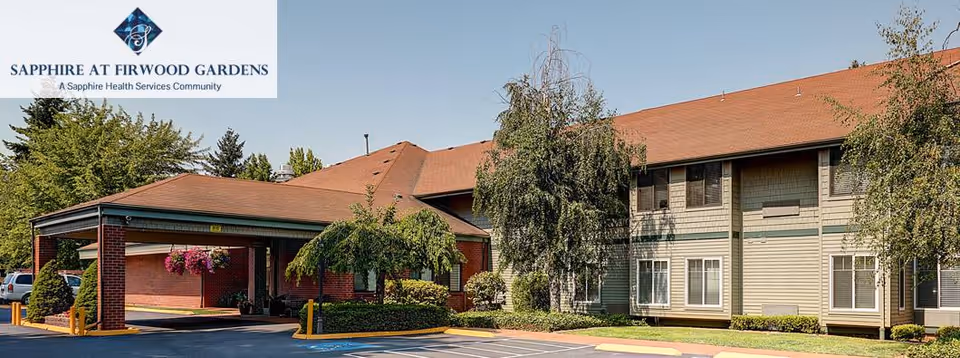 Exterior view of Sapphire at Firwood Gardens, a senior living facility with a covered entrance, brick and siding exterior, surrounded by trees and landscaping under a clear sky.