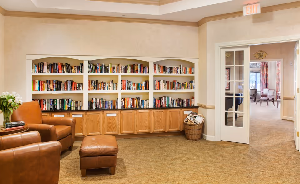 Cozy reading room with built-in bookshelves, leather chairs and an ottoman beside a doorway to another seating area.