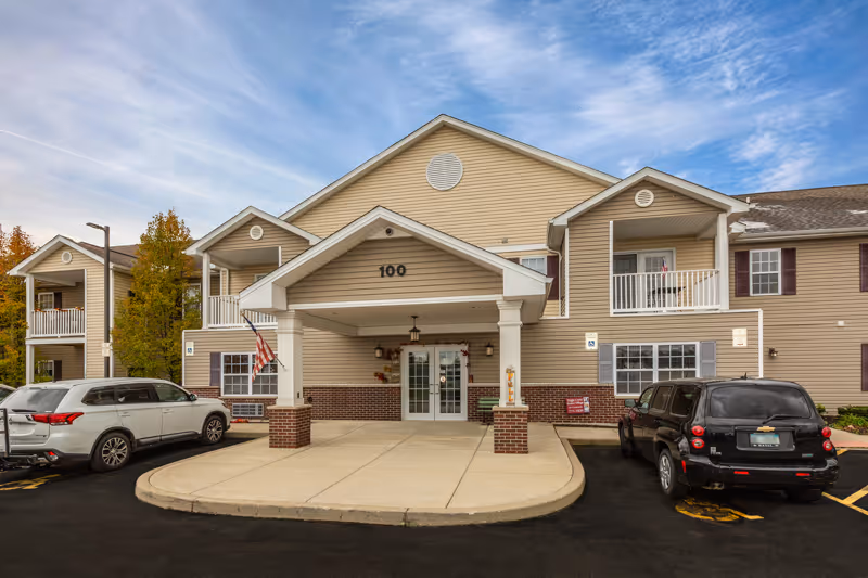 Front exterior view of a beige two-story retirement community building with a covered entrance labeled 100. There are two cars parked in front, an American flag hanging near the entrance, and a clear sky with some clouds above.