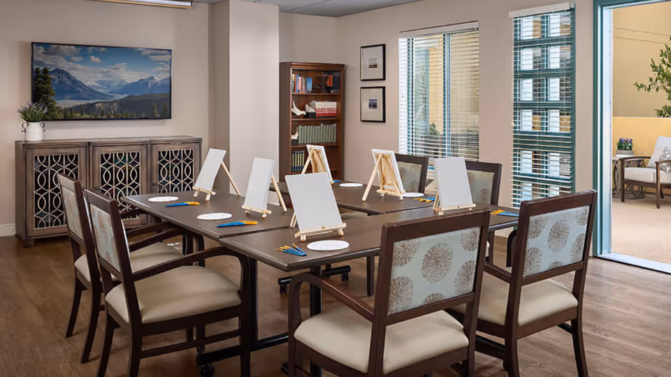 A well-lit activity room with a rectangular table set up for painting, featuring six chairs around the table. Each place has a small easel with a blank canvas, paintbrushes, and paint palettes. The room has wooden flooring, a cabinet with decorative doors, a bookshelf with books, framed pictures on the wall, and large windows with blinds. An open door leads to an outdoor patio area with seating and plants.