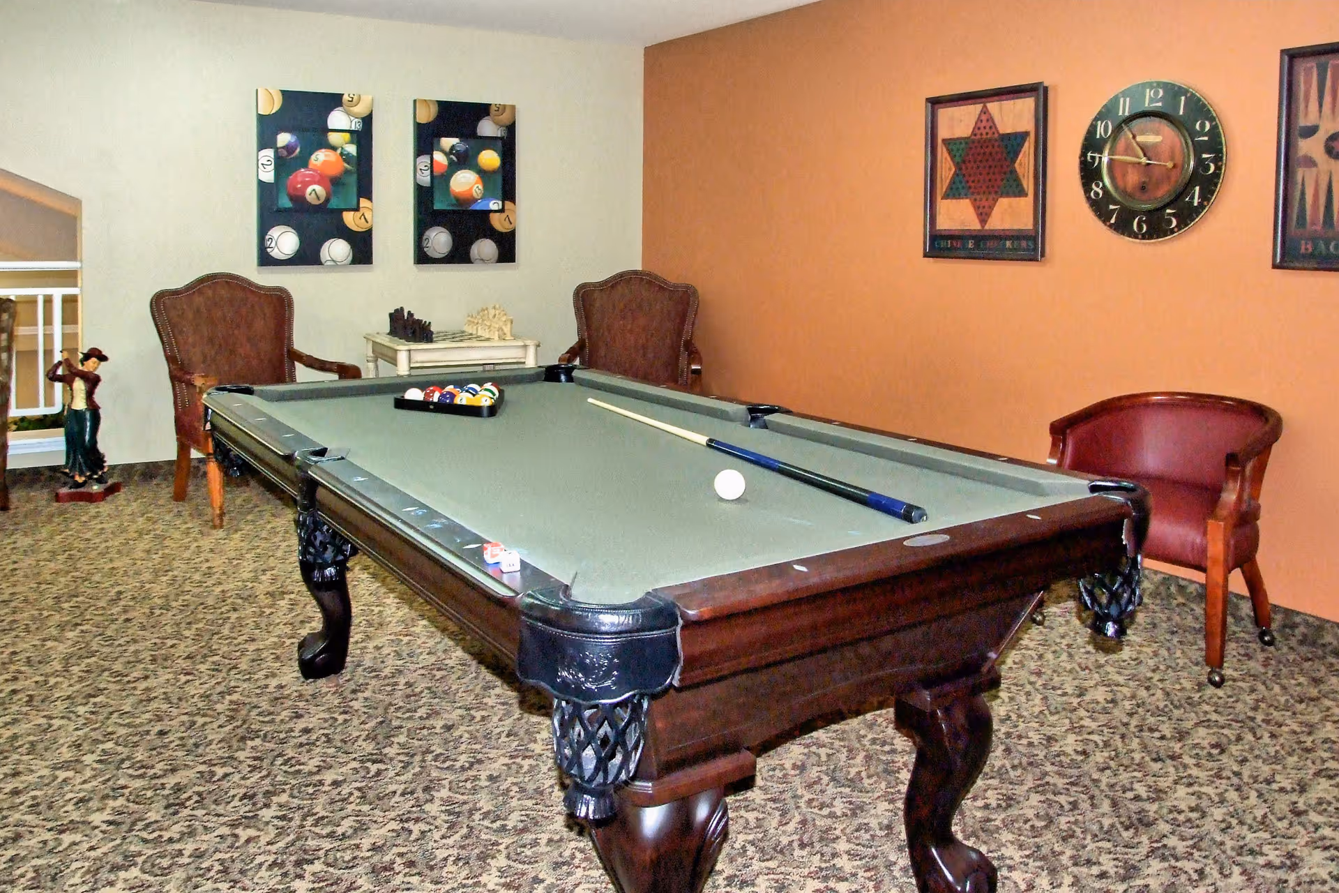 A game room with a pool table in the center, pool balls racked and a cue stick resting on the table. The room has patterned carpet, two upholstered armchairs, a small table with a chess set, and wall decorations including a clock and framed pictures of billiard balls and other designs.