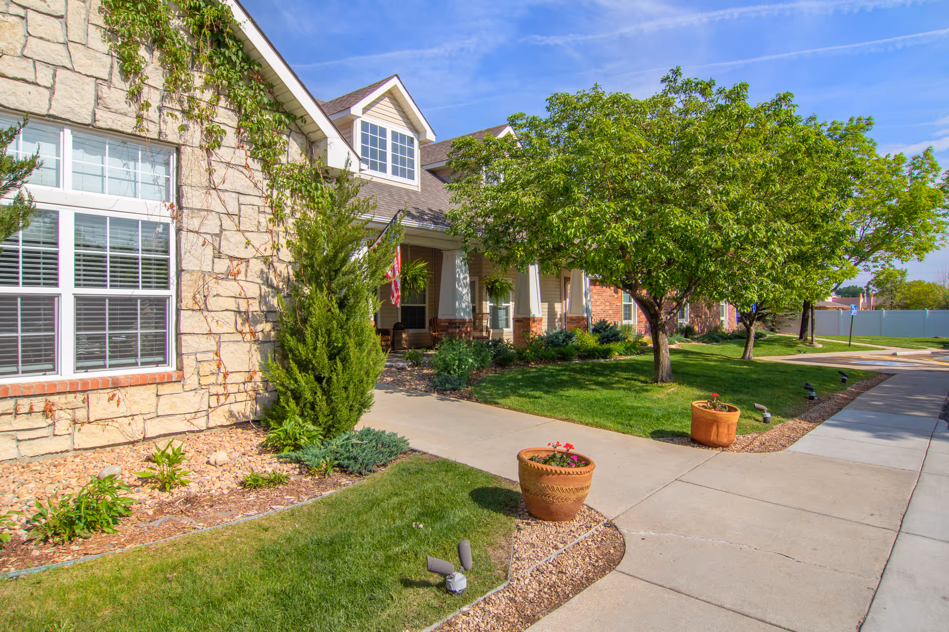 Exterior view of Brookdale Longmont facility showing a stone and brick building with large windows, a covered porch with hanging plants and rocking chairs, a well-maintained lawn, trees, flower pots, and a clear blue sky.