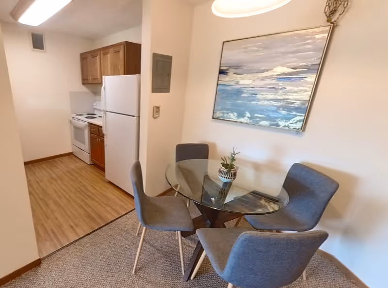 A small dining area with a round glass table and four gray upholstered chairs. A decorative plant is placed on the table. Behind the dining area is a kitchen with wooden cabinets, a white refrigerator, and a white stove. A large abstract painting with blue and white tones hangs on the wall above the dining table.