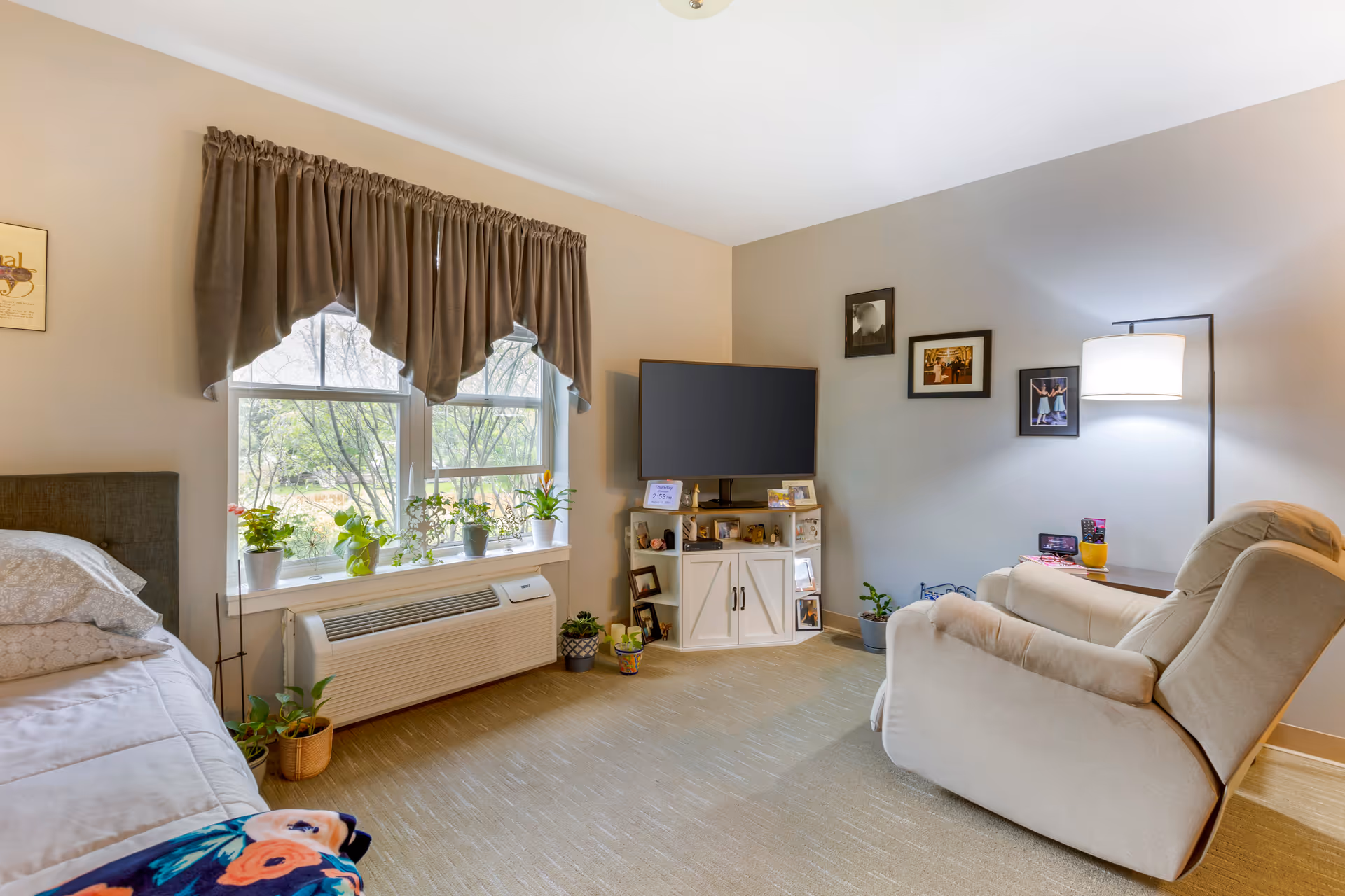 A cozy living space with a beige armchair, a white cabinet holding a flat-screen TV, several framed photos on the wall, and a window with brown curtains and potted plants on the windowsill. The room has beige carpet and neutral-colored walls.