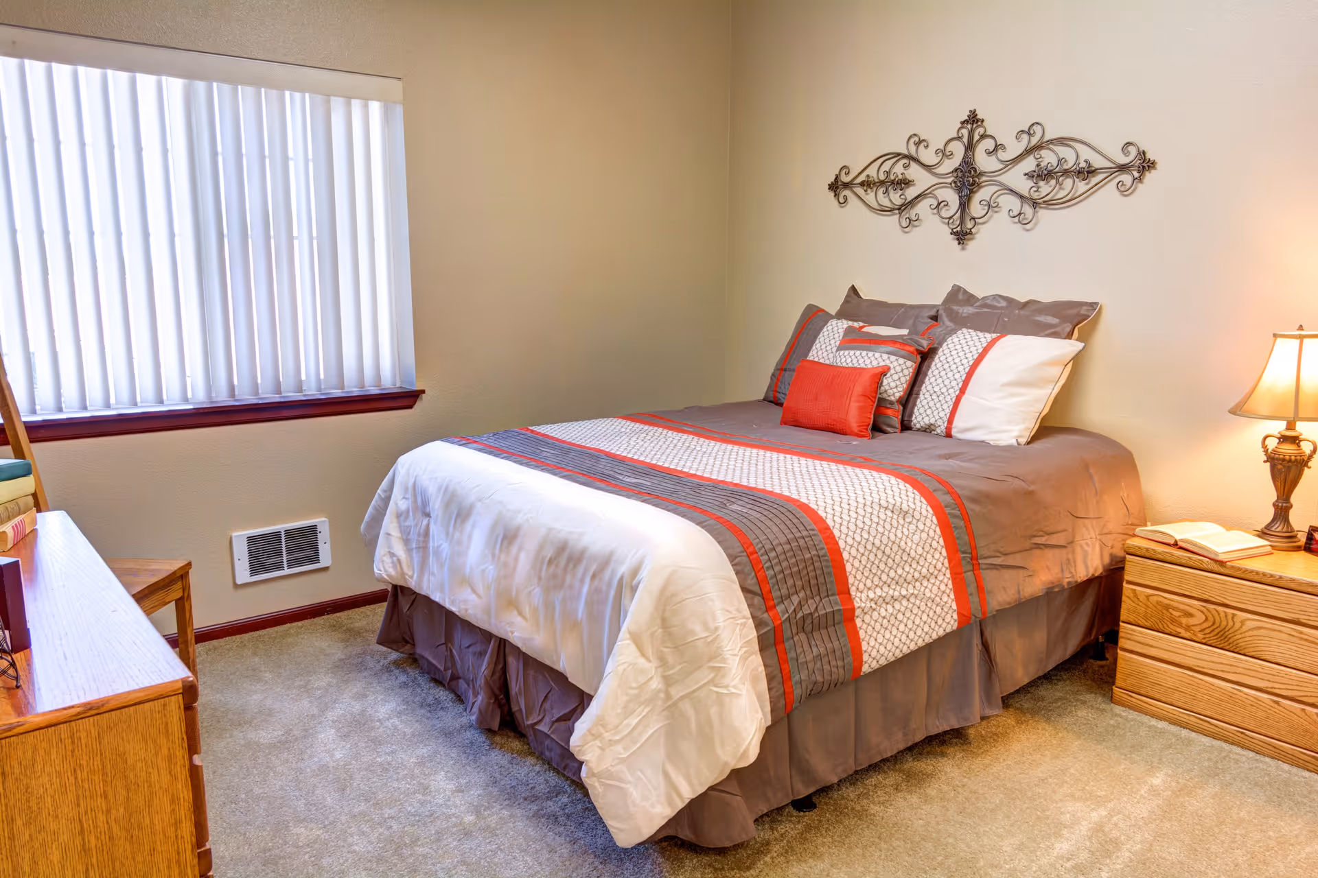 A neatly made bed with a gray, white, and red striped comforter and multiple pillows in a bedroom. There is a wooden nightstand with a lamp and an open book on the right side of the bed. A decorative metal wall art piece hangs above the bed. A window with vertical blinds is on the left wall, and a wooden dresser with books is partially visible.